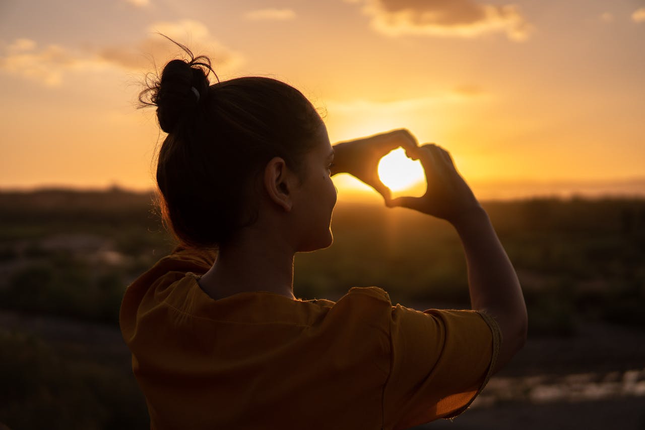"Woman forming a heart with her hands at sunset, symbolizing a way how to be happier through self-compassion, love, and inner happiness."