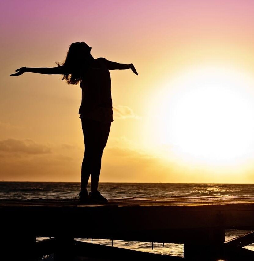 A joyful silhouette of a woman standing on a dock at sunset, embracing the journey of becoming 1% Happier Me Today