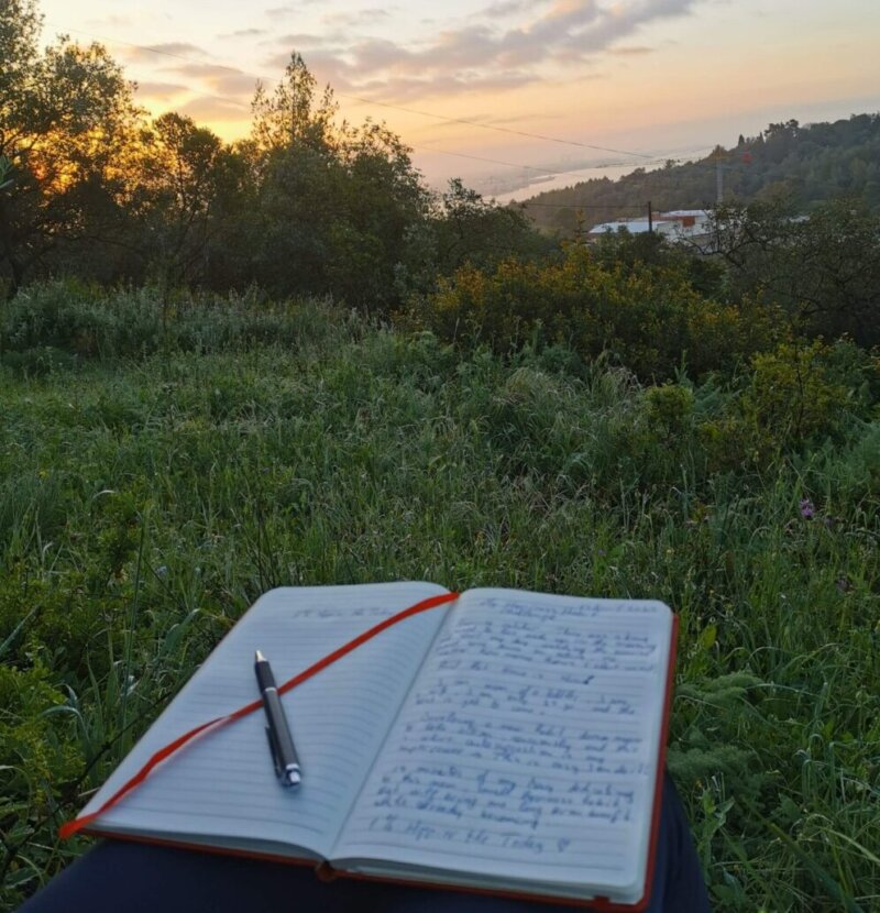 Morning view of sunrise over nature and harbor, with a journal on lap — symbolizing emotional clarity, reflection, and the 1% Happier Me Today daily ritual.