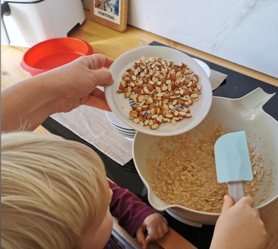 Toddler holding a spatula, assisting parent in preparing a sugar-free banana bread with chopped almonds.