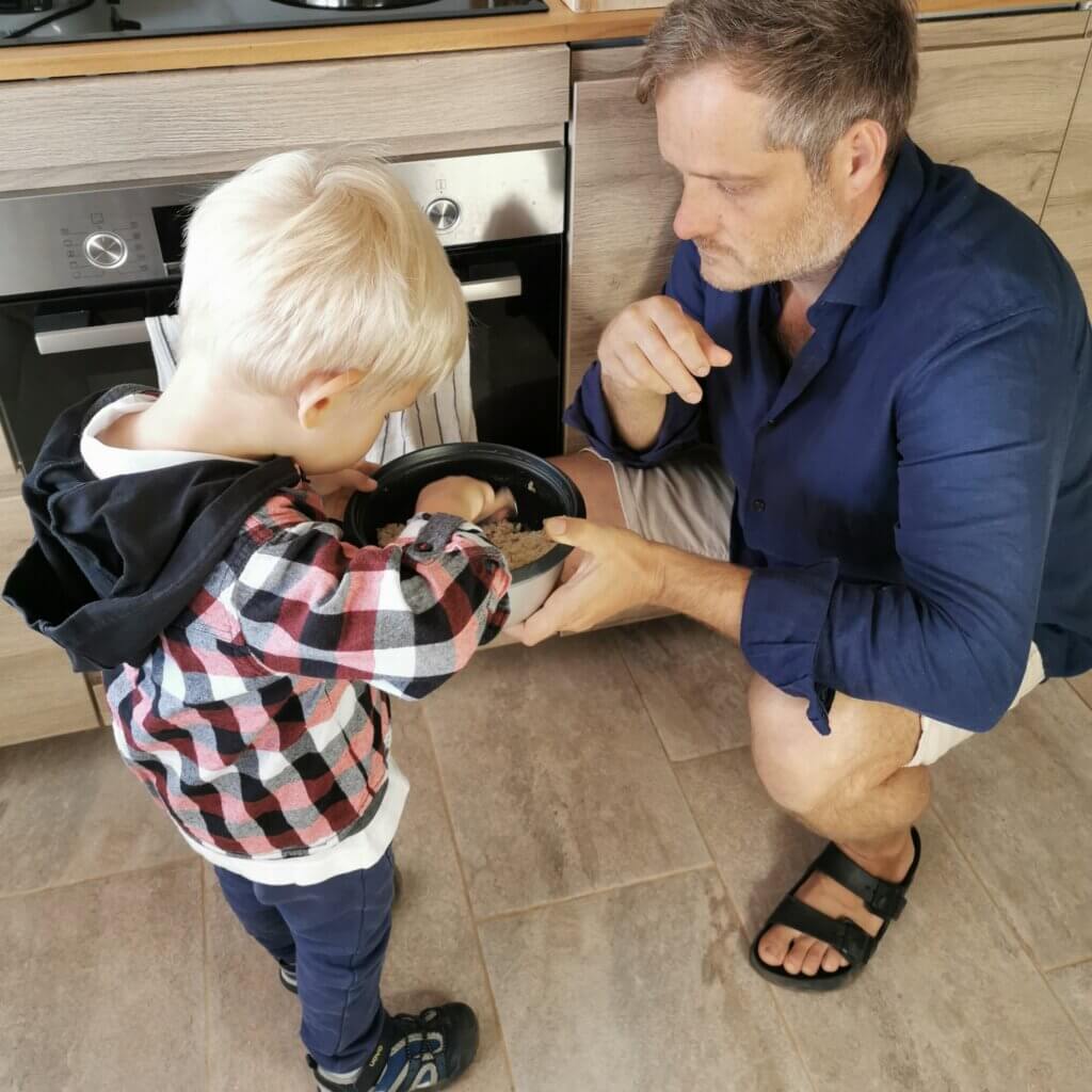 A toddler eats rice from a bowl held by his father, dressed and ready to go out — a candid moment of honouring needs in transition and an example of raising emotionally intelligent kids