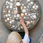 A young child sits proudly beside a handmade umbrella base, decorated with shells, almost completing a family project.
